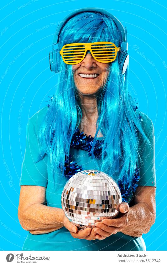 Fröhliche ältere Frau mit blauer Perücke, die mit einer Discokugel und einer Shutter-Brille steht und über Kopfhörer Musik hört, vor blauem Hintergrund