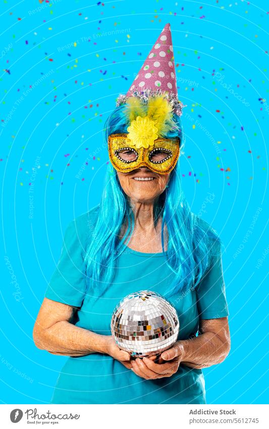 Glückliche ältere Frau, die eine Karnevalsmaske, eine blaue Perücke und einen Partyhut trägt und eine Discokugel in der Hand hält, während sie vor einem blauen Hintergrund im Studio steht