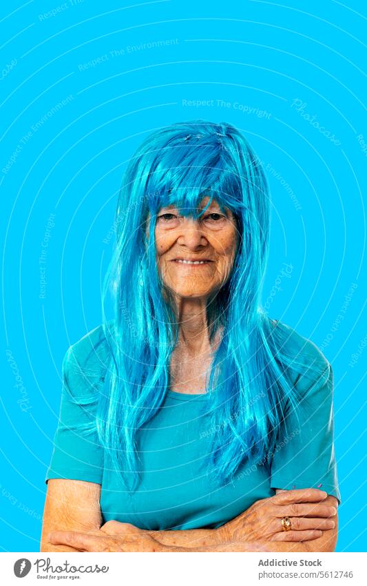 Lächelnde ältere Dame mit blauer Perücke schaut in die Kamera, während sie mit verschränkten Armen auf blauem Hintergrund steht Frau Porträt die Hände gekreuzt