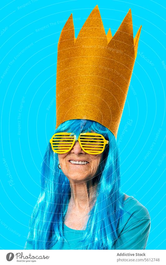 Lustige ältere Frau mit blauer Perücke, goldener Papierkrone und Sonnenbrille, die in die Kamera auf blauem Hintergrund schaut Porträt Krone Streifen