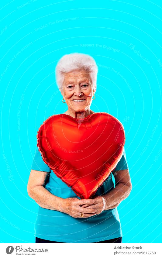Lächelnde ältere Frau, die einen herzförmigen Ballon hält und in die Kamera schaut, vor blauem Hintergrund Liebe Herz Form Luftballon Valentinsgruß