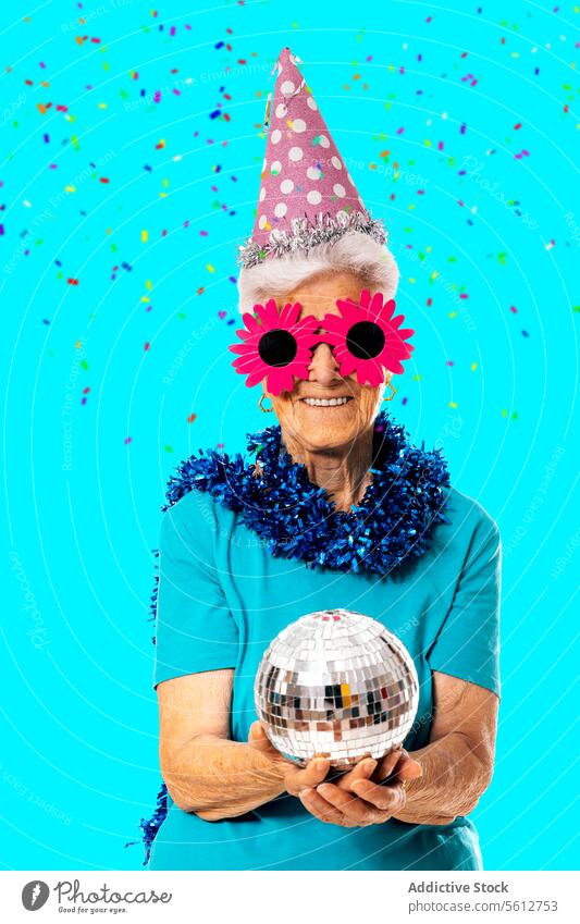 Lächelnde ältere Dame mit Partykappe, lustiger Sonnenbrille und Lametta um den Hals, die eine Discokugel auf blauem Hintergrund hält Frau Ball Neujahr feiern