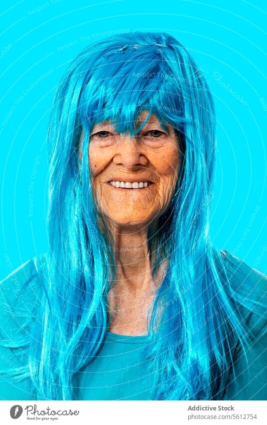 Lächelnde ältere Dame mit blauer Perücke schaut in die Kamera, während sie auf blauem Hintergrund steht Frau Porträt Vorschein in den Ruhestand gehen Hautfalten