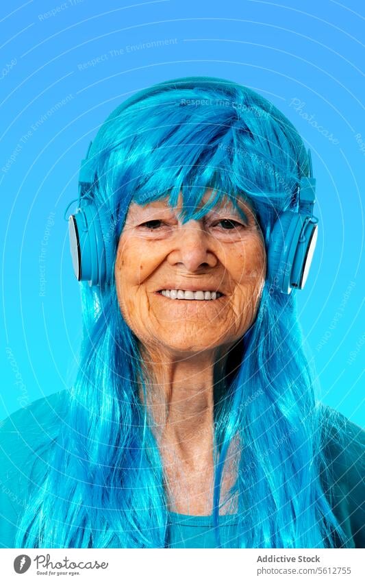 Fröhliche ältere Frau, die über Kopfhörer Musik hört und in die Kamera schaut, vor blauem Hintergrund Porträt zuhören genießen Gesang Perücke Farbe Streichholz