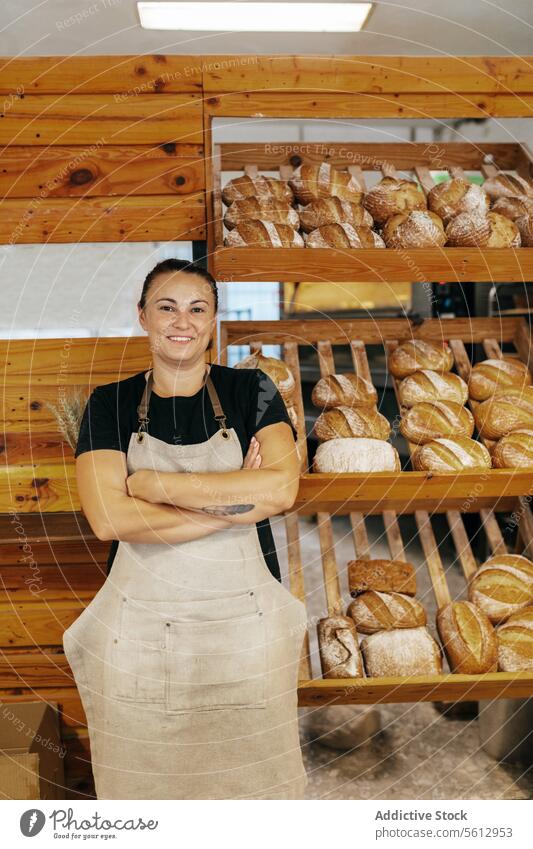 Glückliche Verkäuferin neben Brot in einer Bäckerei Regal Brotlaib selbstbewusst Lächeln die Arme verschränkt Schürze frisch gebacken Lebensmittel hölzern