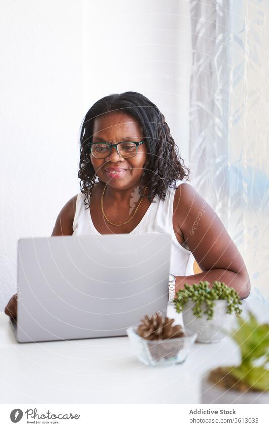 Reife Frau mit drahtlosem Computer zu Hause Laptop Tisch Gardine Pflanze heimwärts Brille Lifestyle heimisch Lächeln Behaarung lockig schwarz Fokus