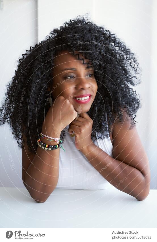 Frau mit Afrofrisur zu Hause Lächeln Wegsehen berühren Kinn Lehnen Porträt weiß Afro-Look Tisch schwarz Frisur heimwärts lockig Lifestyle betrachtend heimisch