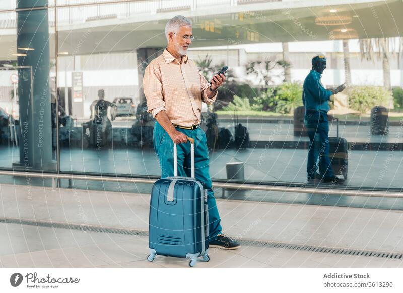Älterer Passagier benutzt Mobiltelefon am Flughafen Senior Mann Nachricht Smartphone Fenster Flugplatz Ganzkörper Brille Vollbart stehen soziale Netzwerke