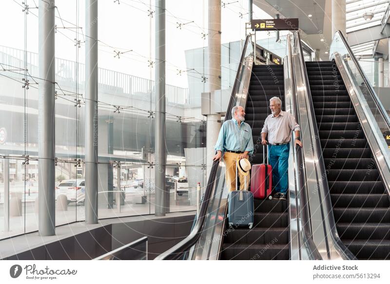 Freunde mit Gepäck auf der Rolltreppe im Flughafen reden Reisender Männer Terminal niedriger Winkel stehen bewegend Treppe Koffer Hut Transit Senior