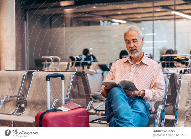 Ein älterer Mann liest ein Buch am Flughafen Passagier lesen Stuhl Vollbart Brille Freizeitkleidung sitzen warten räkeln Hobby lernen Fokus ernst Konzentration