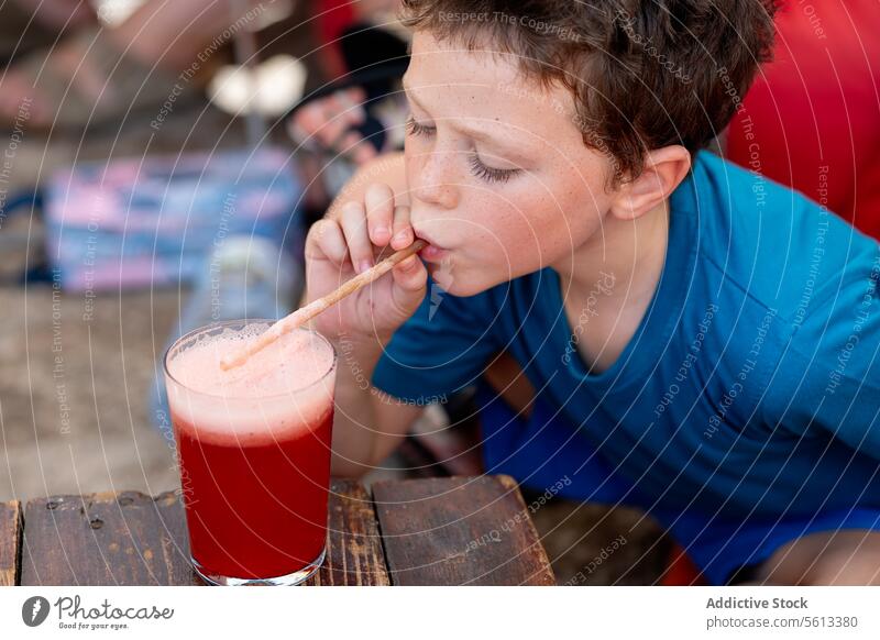 Hoher Blickwinkel auf kleinen Jungen in Freizeitkleidung, der frischen Wassermelonensaft in einem Glas mit Strohhalm trinkt, während er an einem Holztisch im Restaurant sitzt
