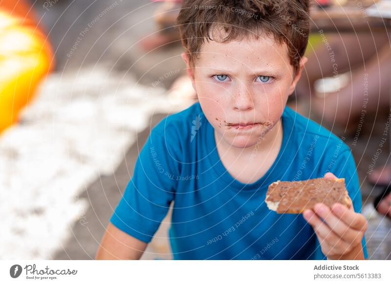 Süßer hungriger Junge in Freizeitkleidung schaut in die Kamera, während er süße Schokolade mit Brot zum Frühstück isst, während er im Restaurant sitzt essen