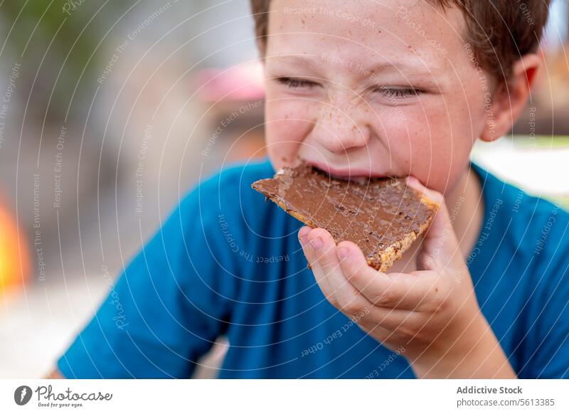 Süßer hungriger Junge in Freizeitkleidung mit geschlossenen Augen isst süße Schokolade mit Brot zum Frühstück, während er im Restaurant sitzt essen Aufstrich