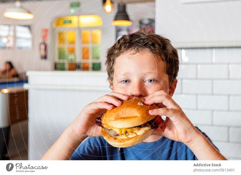 Porträt eines hungrigen Jungen im Grundschulalter, der einen leckeren Burger isst und in die Kamera schaut, während er seine Freizeit in einem Restaurant am Wochenende verbringt