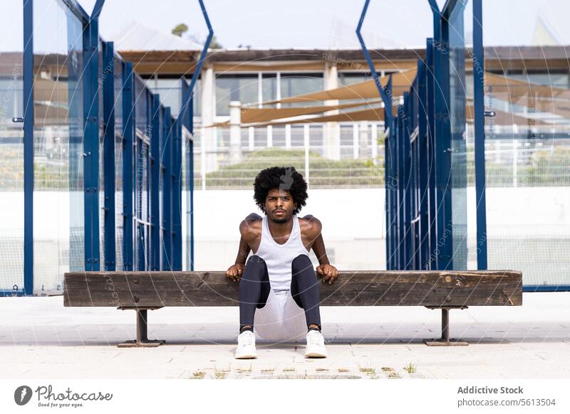 Selbstbewusster, ernster afroamerikanischer männlicher Sportler, der seinen Körper auf einer Holzbank balanciert und eine Kniebeuge macht, während er in die Kamera in der Stadt schaut