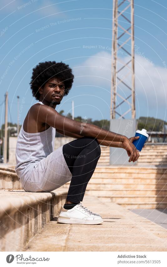 Seitenansicht eines afroamerikanischen müden männlichen Sportlers in Sportkleidung, der auf einer Treppe sitzt und eine Flasche Wasser bei einer Trainingspause an einem sonnigen Tag trinkt