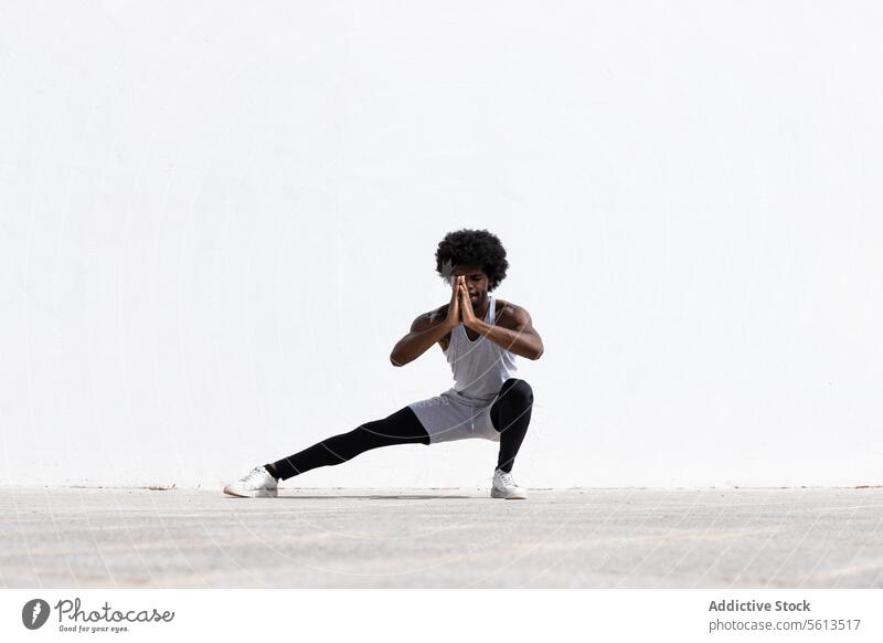 Ernster schwarzer Sportler, der über einer weißen Wand trainiert Athlet Mann Übung Afrohaar Gebet Pose Dehnung Bein Kniebeuge Gleichgewicht Straße Ausdauer