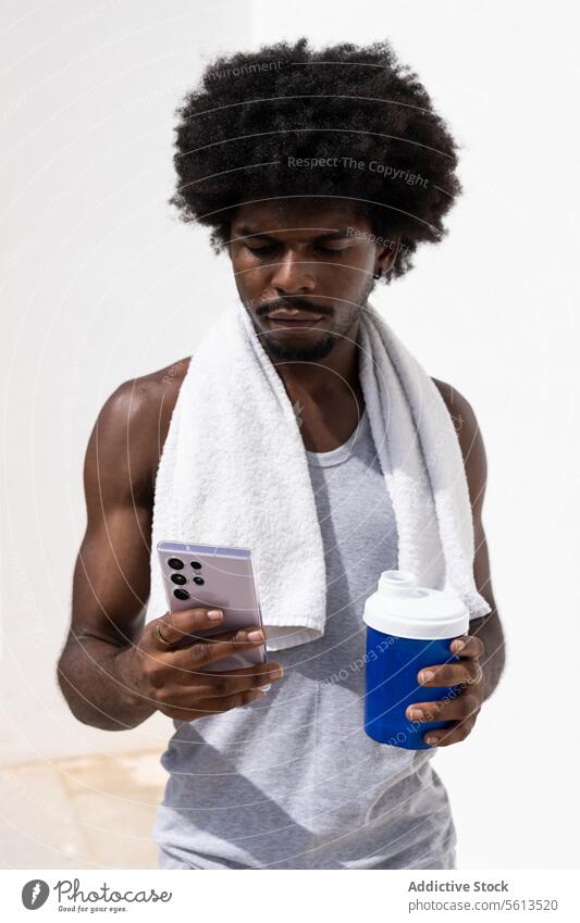 Afroamerikanischer junger Sportler mit Afrohaar und Handtuch im Nacken, der auf den Bildschirm schaut, während er auf seinem Handy surft und eine Flasche Wasser an eine weiße Wand hält
