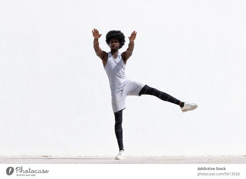 Ernster schwarzer Mann streckt Körper über weiße Wand Athlet Übung Afrohaar Dehnung Arme selbstbewusst Fokus Gleichgewicht anstrengen Sportler ernst Bein stehen