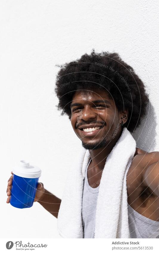 Lächelnder junger männlicher Sportler mit Afro-Haar, der eine Wasserflasche hält und ein Selfie macht, während er sich nach dem Training im Sommer an eine weiße Wand lehnt