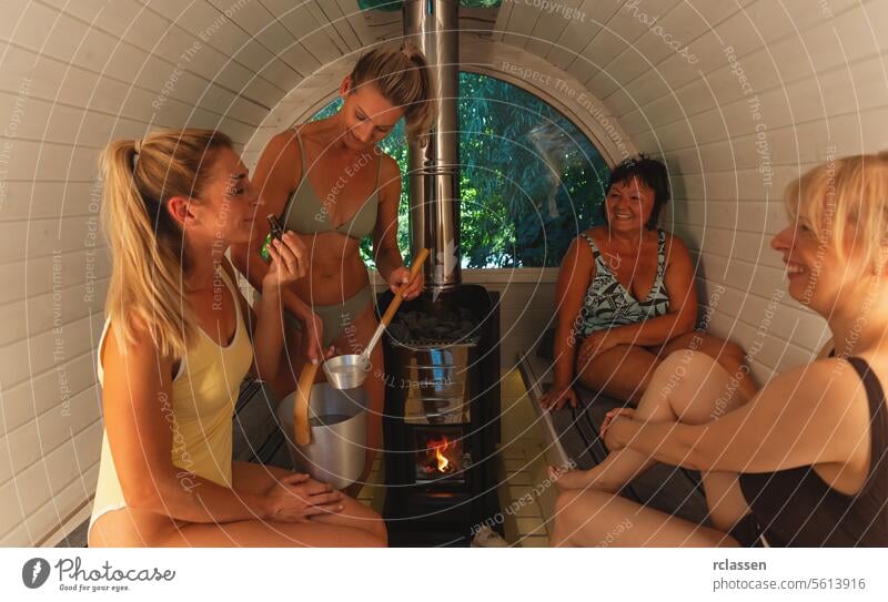 Frauen in einer Sauna, eine hält eine Flasche mit Saunaduft in der Hand und riecht daran, die anderen lächeln tropfend ätherisches Öl Wasser gießen heiße Steine