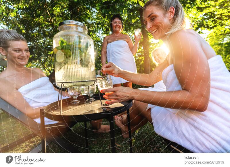 Frauen in Badetüchern, die nach der finnischen Sauna in einem Garten mit Limetten aufgegossenes Wasser aus einem Spender servieren. Finnisch Zitrone Kalk