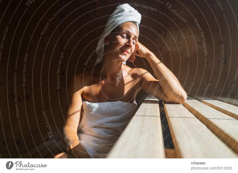 Frau in einer finnischen Sauna, zurückgelehnt, Augen geschlossen, mit einem Handtuch um den Kopf gewickelt, im Sonnenlicht eines Kurortes vihta Wellnessbad
