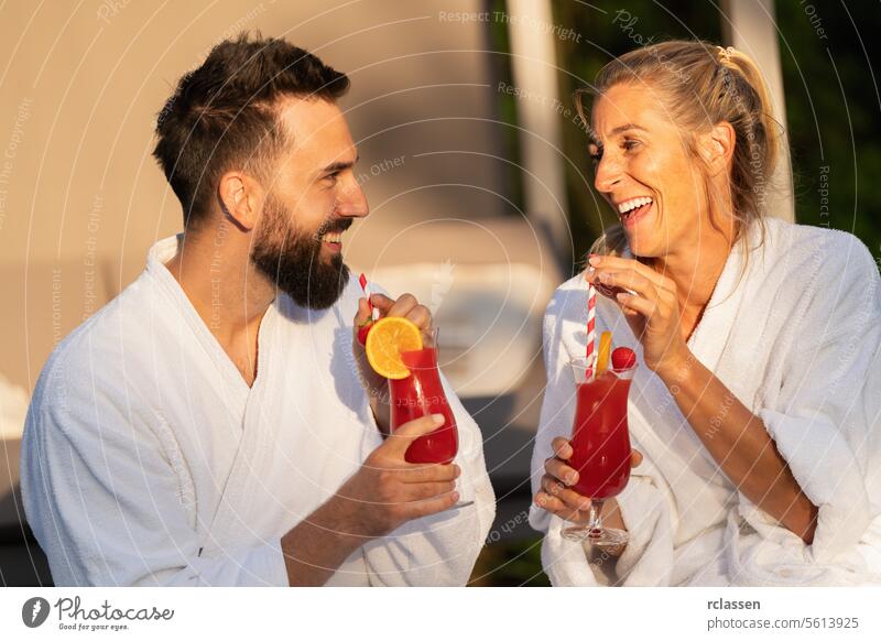 Zwei Menschen in weißen Bademänteln lachen und trinken Cocktails im Sonnenlicht in einem Wellness-Hotel schlürfend Bademantel Spa-Wellness-Resort Paar