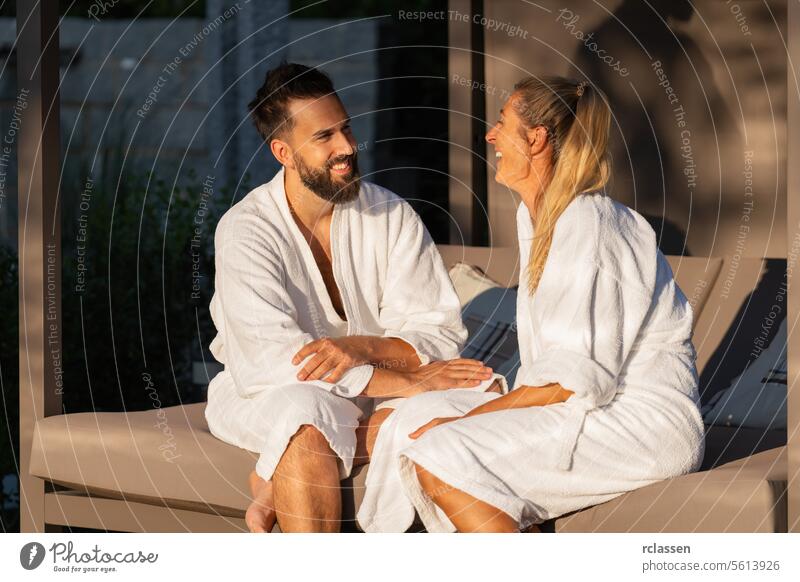 glückliches Paar in weißen Bademänteln, das auf einer Liege im Freien im Sonnenlicht im Spa-Wellness-Hotel sitzt und spricht Bankliege sprechend Lächeln