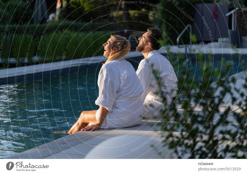 Paar in weißen Bademänteln am Pool sitzend, nach oben schauend und lachend, ein Tag in einem Wellness-Hotel Wellnessbad Resort weiße Gewänder aufschauend