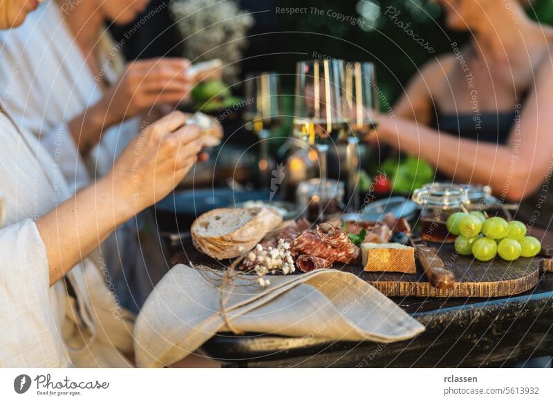 Nahaufnahme eines Tisches mit Weingläsern, Brot und Weintrauben, im Hintergrund speisen Freunde speisend Trauben Charcuterie Outdoor-Essen Abend Käse Mahlzeit