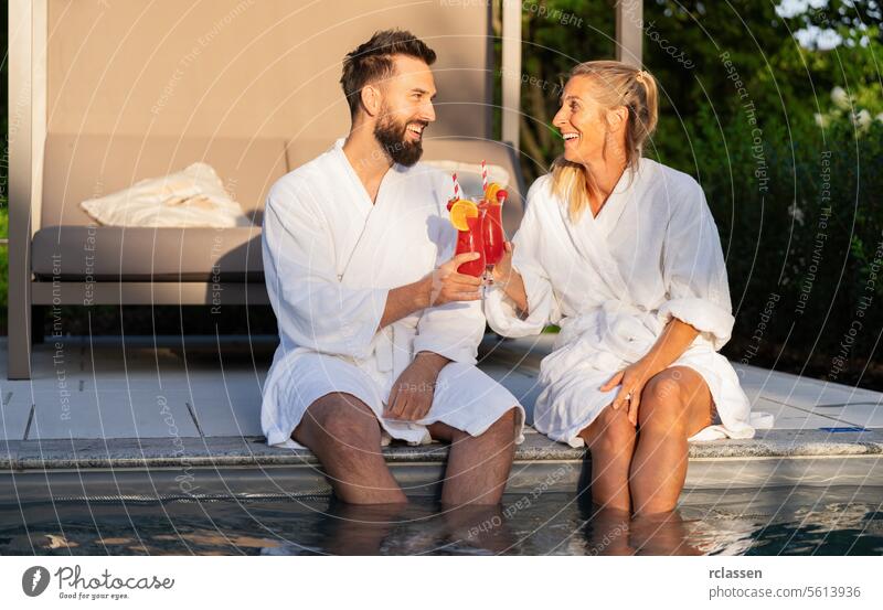 Zwei Menschen in weißen Bademänteln lachen und stoßen mit roten Getränken am Pool eines Wellnesshotels auf den Sonnenuntergang an Spa Erholung Cocktails Freunde