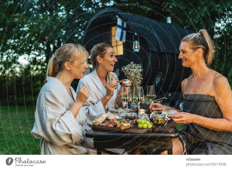 Freunde in Bademänteln genießen eine Mahlzeit und Wein in der Nähe eines finnischen Saunafasses im Freien Nebel Fahne finnische Sauna Genuss speisend Erholung