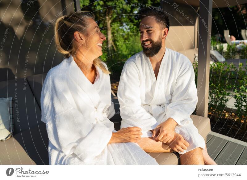 Lächelndes Paar in weißen Bademänteln genießt einen sonnigen Tag im Freien auf einer Paar-Liege in einem Wellness-Hotel Liebe zusammen lachend Resort
