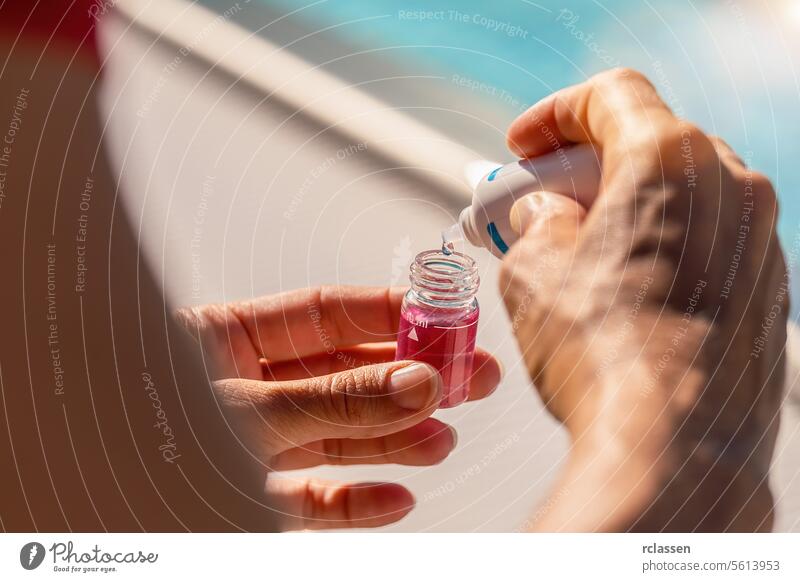 Hände eines Schwimmbadtechnikers, der mit einer Tropfflasche Lösungsmittel in ein Fläschchen mit rosafarbener Lösung für einen pH-Test einfüllt