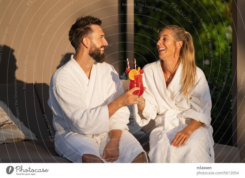 Lächelndes Paar in weißen Bademänteln stößt in sonnigem Ambiente im Kurort auf Cocktails an Gläserklirren Bademantel Wellness Hotel Spa-Wellness-Resort