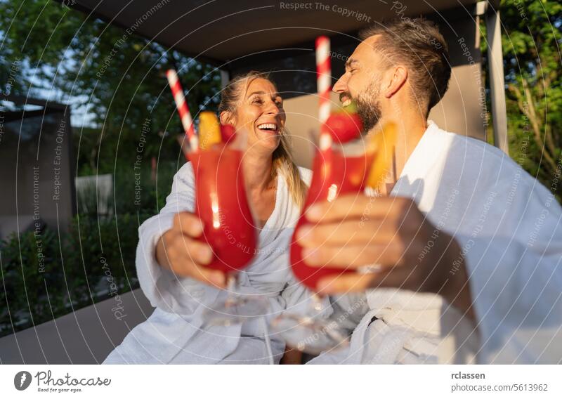 Paar in weißen Gewändern stößt mit roten Cocktails im Freien eines Kurortes an Bademantel Wellness Hotel Spa-Wellness-Resort weiße Gewänder rote Cocktails