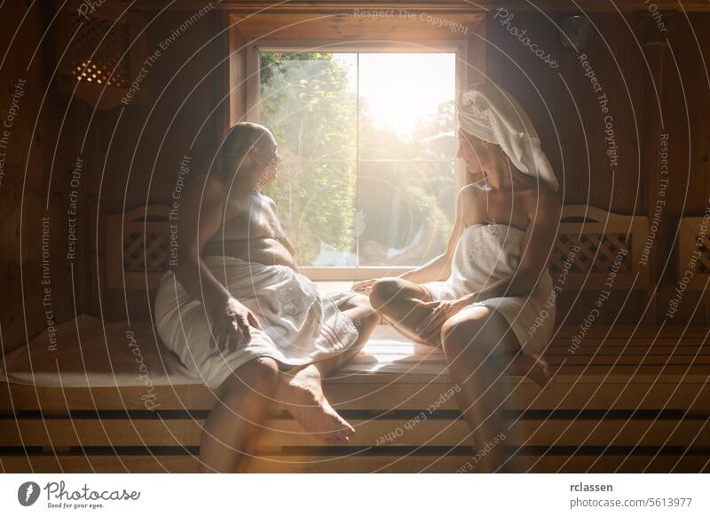 Mann und Frau sitzen sich in einer finnischen Sauna gegenüber, beide in Handtücher gewickelt, Fenster mit Sonnenlicht in einem Wellness-Hotel Schweiß Deutsch