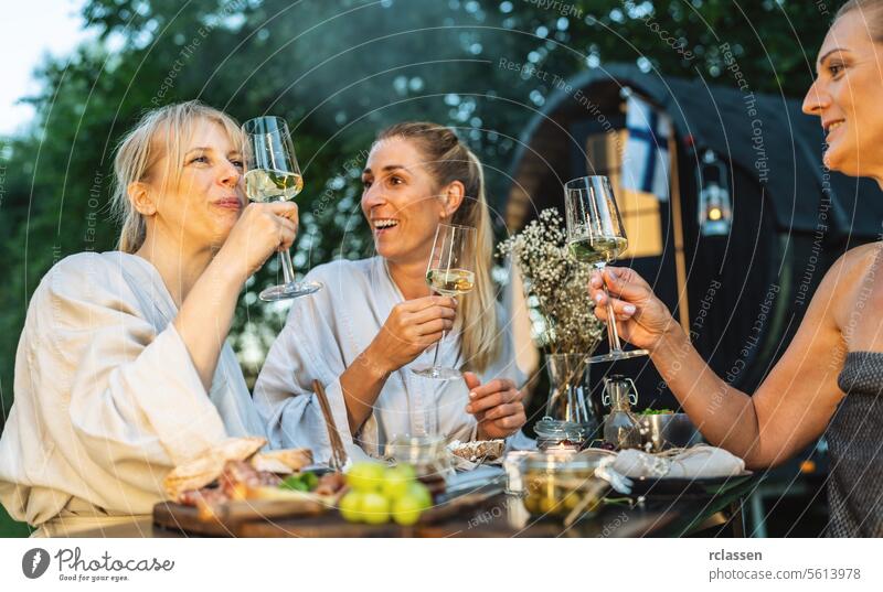Frauen in Bademänteln genießen Wein im Freien, mit einer mobilen finnischen Saunakabine im Hintergrund mobile Sauna Erholung Freunde Genuss Freizeit Natur