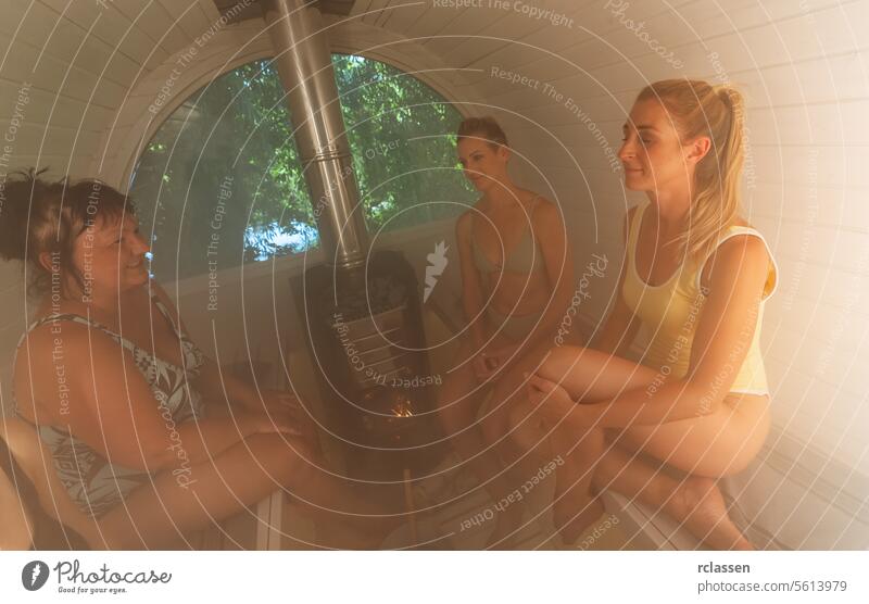 Vier Frauen sitzen in einer finnischen Fasssauna, lächeln und genießen die Hitze Mobile Verdunstung heiße Steine Erholung Wellness Badeanzüge