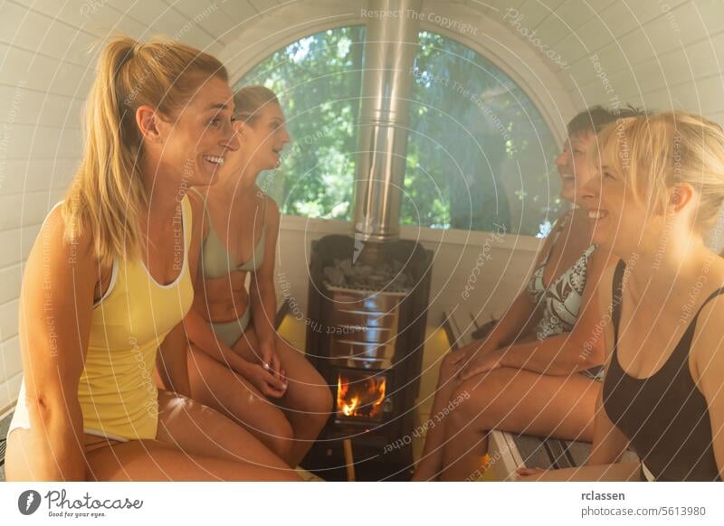 Vier Frauen unterhalten sich in einer hell erleuchteten finnischen Fasssauna mit Ofen. Finnische Fass-Sauna Gespräch Saunaofen Genuss Wellness Gesundheit