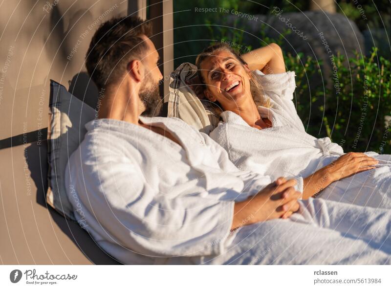 Paar in weißen Bademänteln entspannt und lacht zusammen auf einer Liege im Sonnenlicht in einem Wellness-Hotel Bankliege sich[Akk] entspannen Resort Liebe