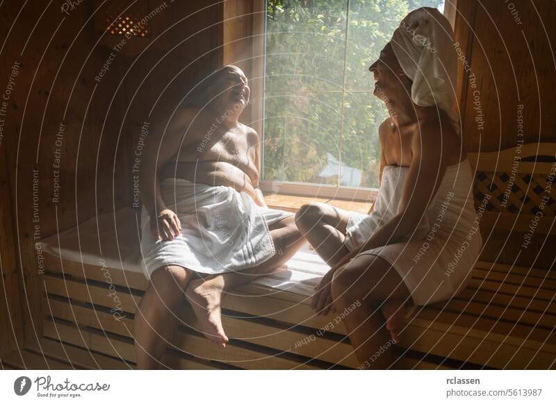 Mann und Frau in einer finnischen Sauna, zusammen lachend, Frau mit Handtuch auf dem Kopf, beide im Wellnesshotel sitzend Wellnessbad Resort Hotel Fenster