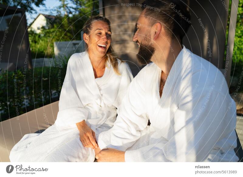 Zwei Menschen in weißen Gewändern sitzen auf einer Paarliege im Freien und lachen zusammen in einem Hotel Liege Liebe zusammen lachend Resort Wellnessbad