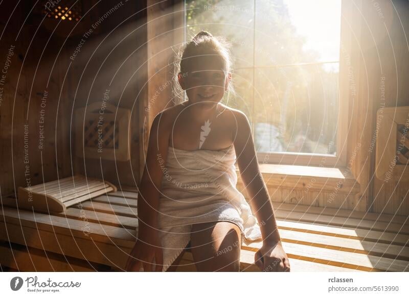 Lächelndes Kind in einer finnischen Sauna, Sonnenstrahlen strahlen durch das Fenster, eingewickelt in ein Handtuch im Spa-Wellness-Hotel Bademantel