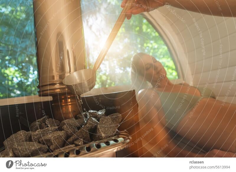 Frau gießt Wasser auf Saunasteine, eine andere Frau entspannt sich im Hintergrund. Mobile Finish Sauna Konzept Bild Wasser gießen heiße Steine Erholung Wellness