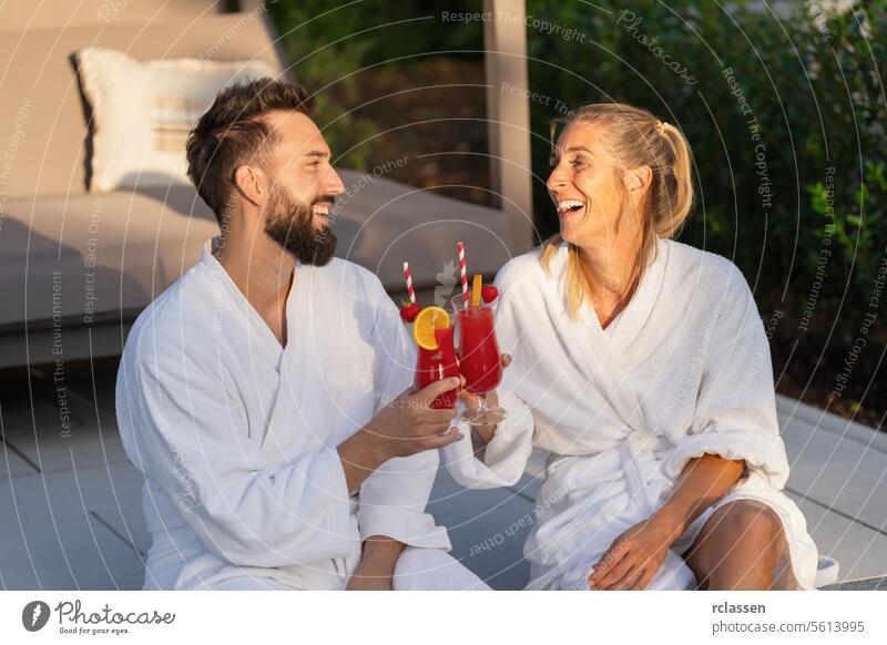 Fröhliches Paar in weißen Bademänteln stößt mit roten Getränken an, im Freien zur goldenen Stunde in einem Hotel schlürfend Bademantel Wellness