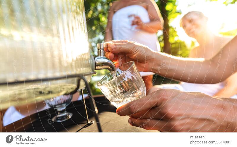 Nahaufnahme einer Hand, die Wasser aus einem Spender gießt, mit Menschen im Hintergrund Finnisch Sauna Erschöpfung Kalk Zitrone Wasser gießen Getränkespender