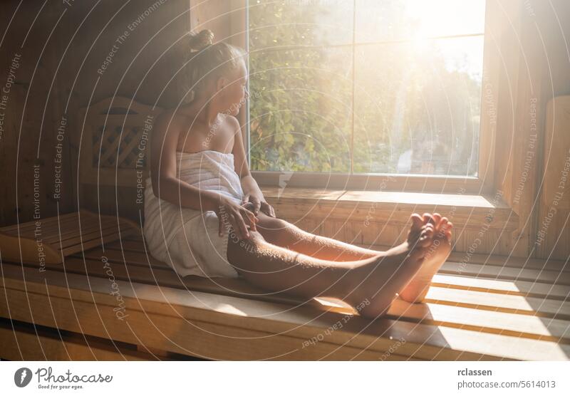 Kind auf einer finnischen Saunabank sitzend, in ein Handtuch eingewickelt, mit Blick aus einem sonnenbeschienenen Fenster in einem Wellness-Hotel Bademantel
