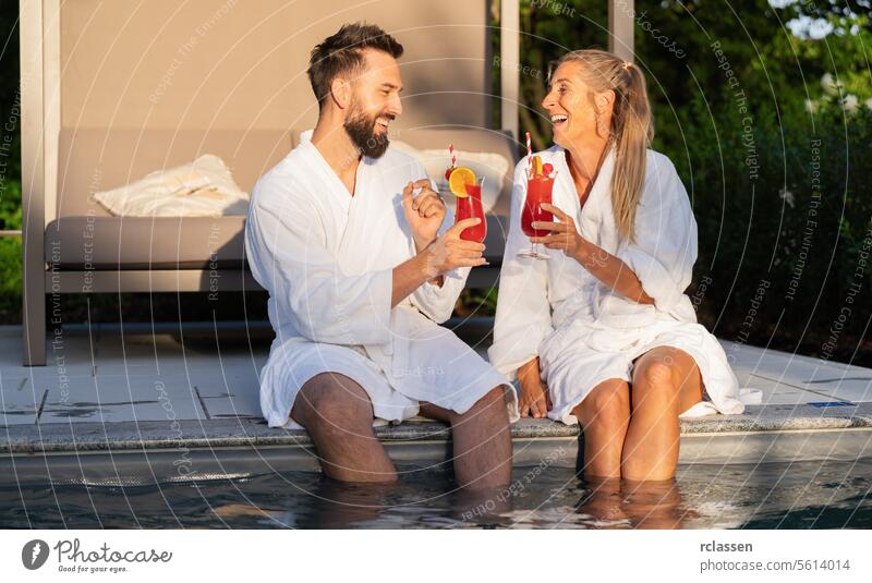Zwei Personen in weißen Bademänteln stoßen mit Cocktails am Pool an und genießen den Sonnenuntergang Spa Erholung Freunde Zuprosten lachen Gläserklirren